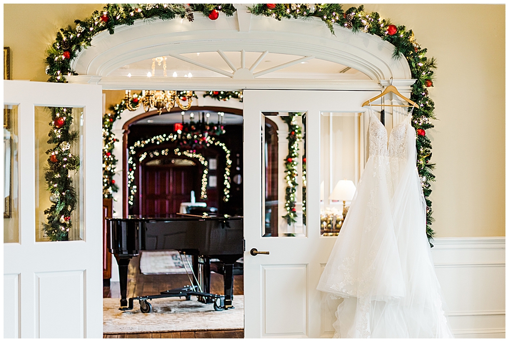 Ballgown lace wedding dress from Zakaa Couture in Ashburn, VA. Hanging on a white french door with Christmas garland around the frame. In the background, a black baby grand piano.