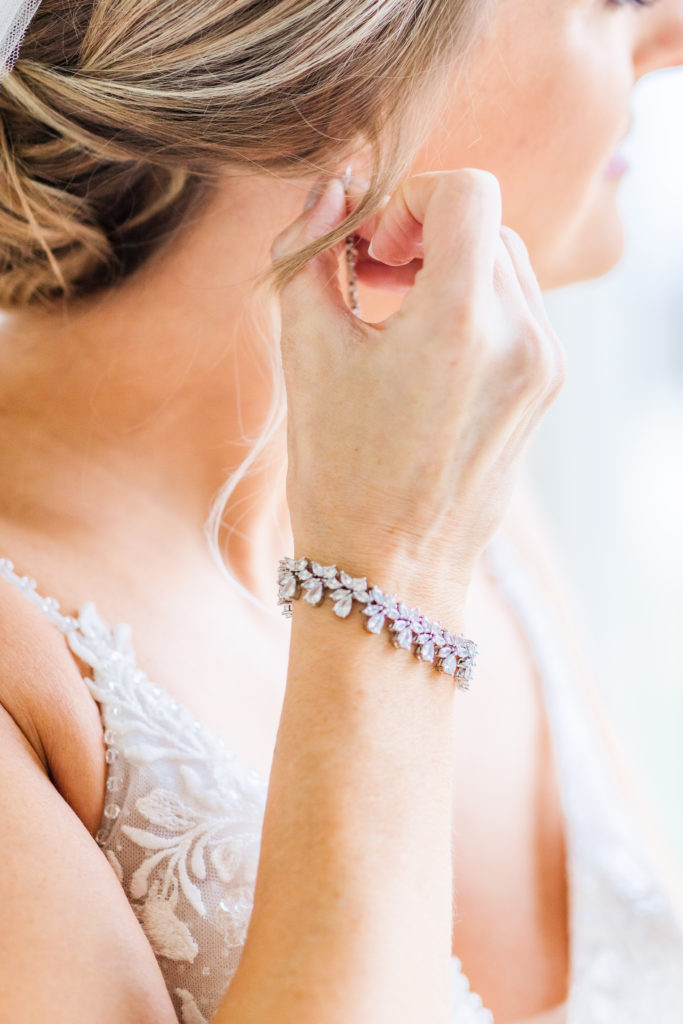 Bride putting on her earrings detail shot, fine art wedding photos. Crystal bracelet and spaghetti strap wedding dress.