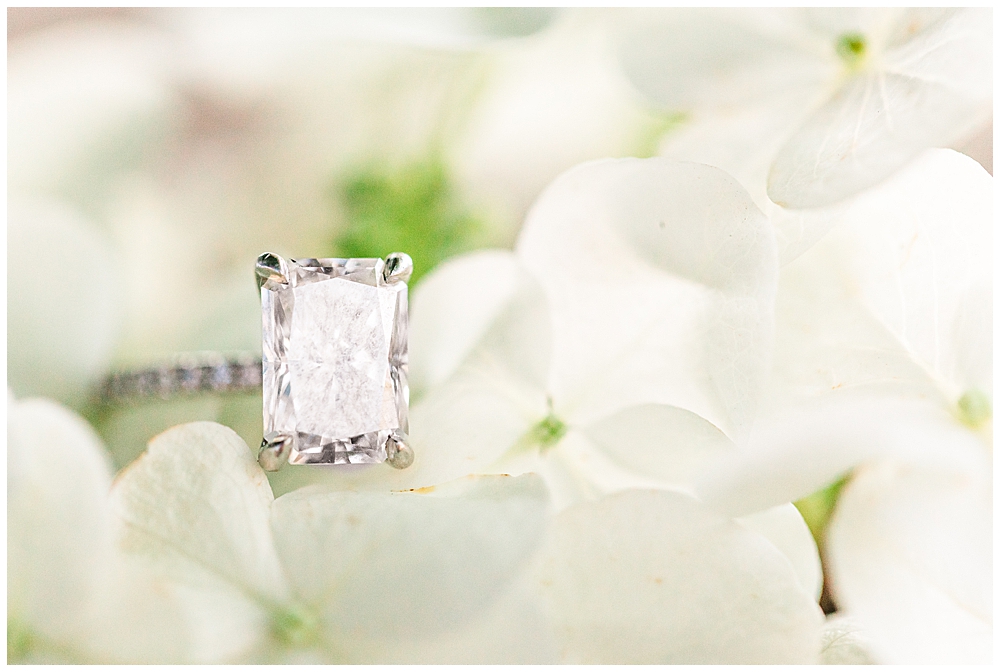Ring shot on white hydrangea flowers, emerald-cut diamond with diamond band and hidden halo band.