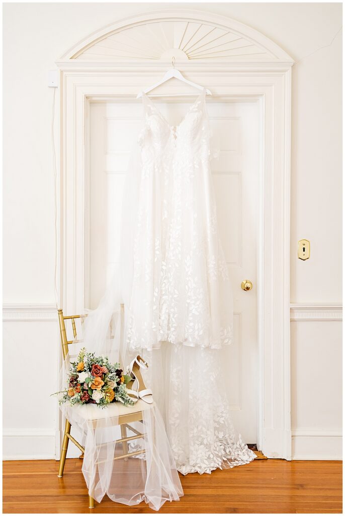 A full length shot of an A-line wedding dress with elegant floral lace details and whimsical off-shoulder straps, and a gold chair in front of it holding a fall colored bouquet of sola wood flowers and the Bride's simple white block-heeled sandals

Rust Manor House Wedding | Leesburg Wedding Photography | VA Wedding Venues