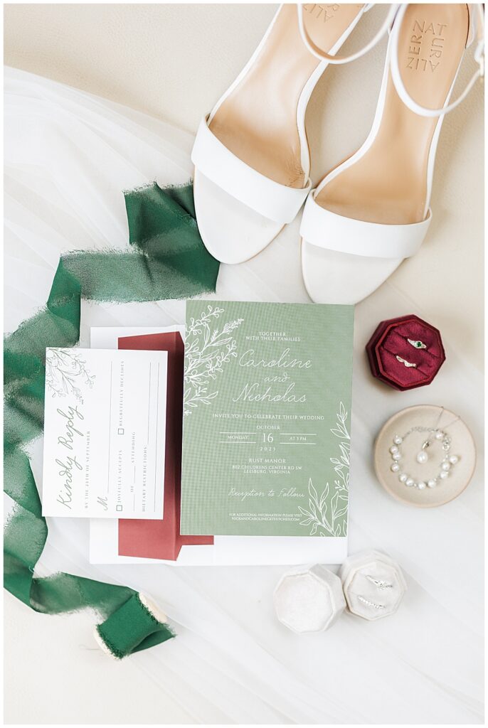 A flat lay shot of an simple two-pieced wedding invitation suite, including a green main invitation card and white and green RSVP card. Around the invitation are the Bride's simple white wedding shoes, a plain tulle veil, a burgundy ring box and pearl jewelry

Rust Manor Wedding | Leesburg Wedding Photography | VA Wedding Venues