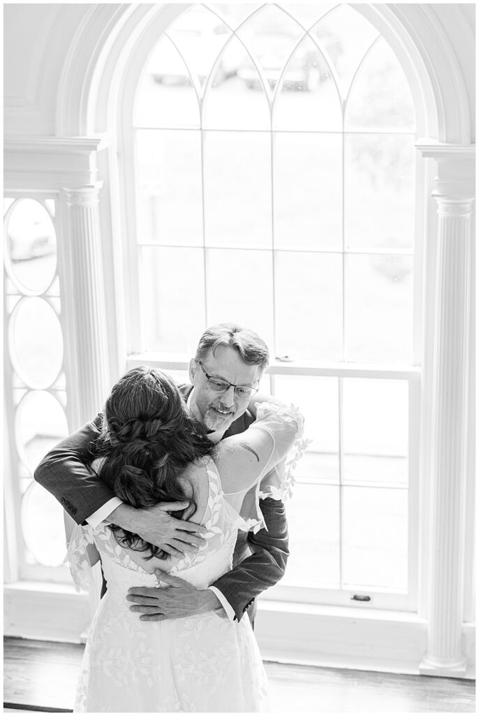 A black and white photo of a father hugging his daughter at the First Look of her in her wedding dress. This bride and father of the bride moment is captured in front of a large curved window. 

Rust Manor House Wedding | Leesburg Wedding Photography | VA Wedding Venues