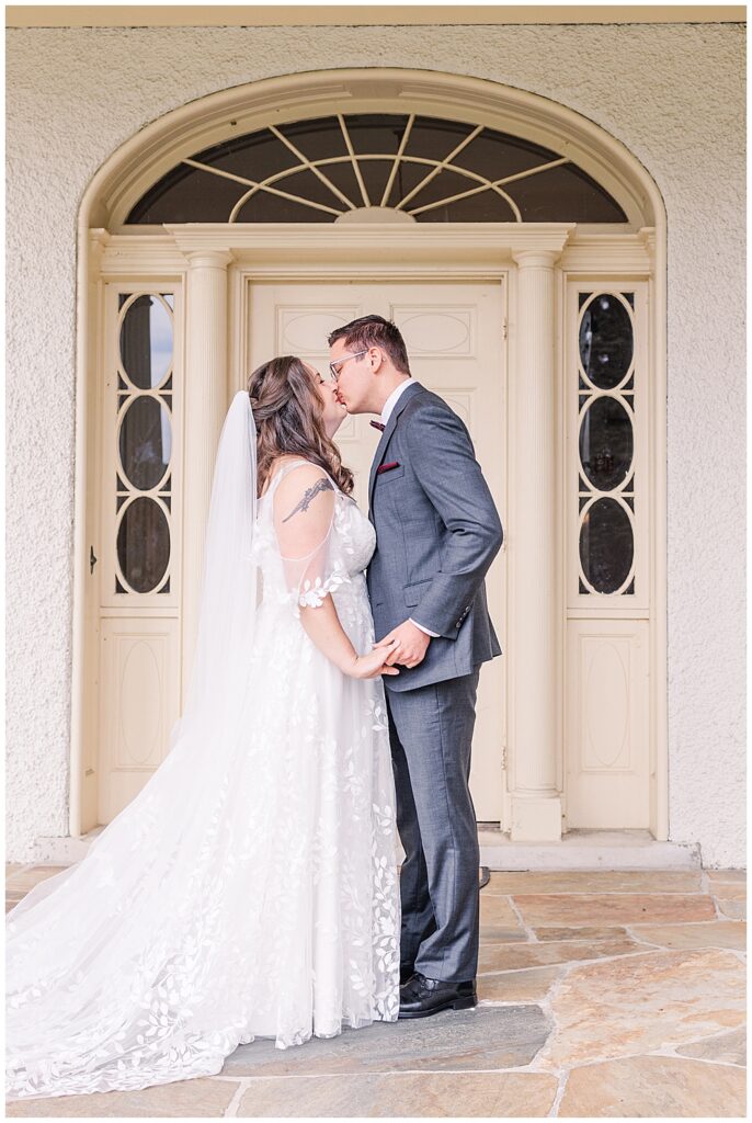 A bride and groom share a kiss right after having their First Look on their wedding day. The groom is wearing a grey 3-piece suit with a burgundy bowtie and pocket square. The bride is in an off-the-shoulder A-line lace gown with a whimsical leafy pattern down the skirt.

Rust Manor House Wedding | Leesburg Wedding Photography | VA Wedding Venues