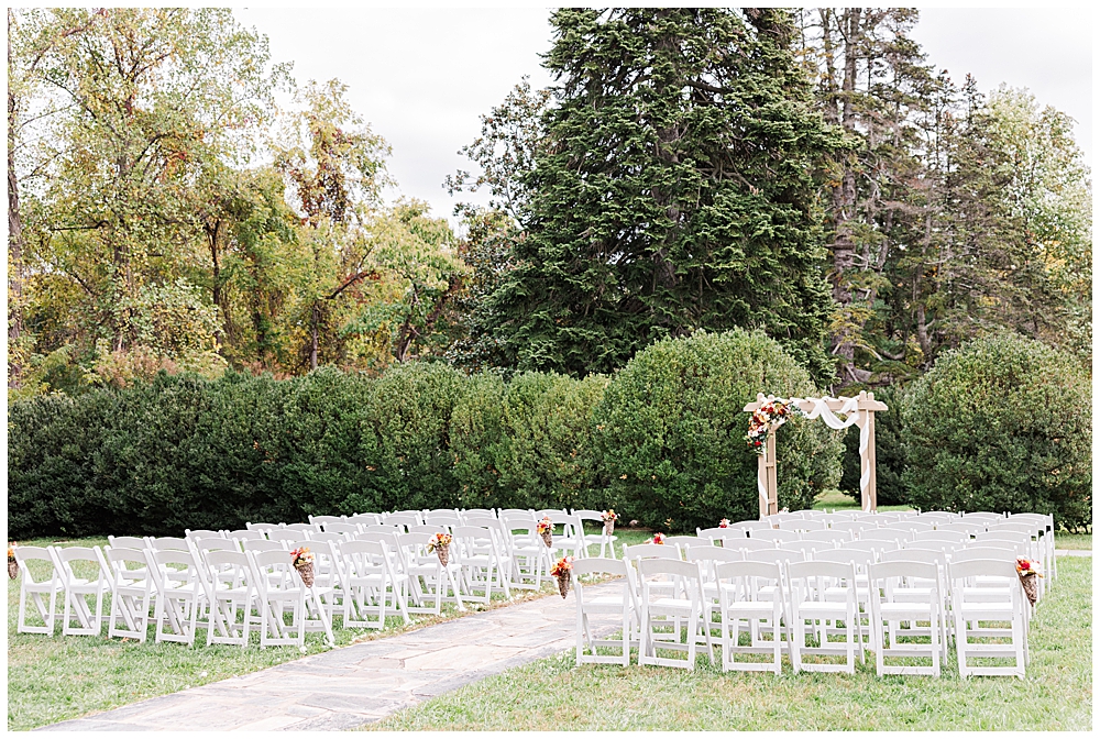 An outdoor ceremony setup with a wooden arbor decorated with loose white fabrics and autumn florals. The white folding chairs are lined up in rows of six with autumn florals on each end.

Rust Manor House Wedding | Leesburg Wedding Photography | VA Wedding Venues