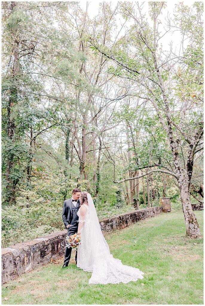 A full-length shot of a bride and groom resting foreheads together for a photo with the bride's long veil and trail fanned out behind her. They're standing next to a low stone wall in the midst of a treelined landscape.

Rust Manor House Wedding | Leesburg Wedding Photography | VA Wedding Venues