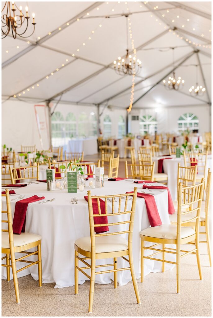 A tented reception setup with round tables adorned in white table cloths, maroon napkins, greenery centerpieces, surrounded by gold padded chairs. The ceiling of the tent is hung with string lights and chandeliers.

Rust Manor House Wedding | Leesburg Wedding Photography | VA Wedding Venues