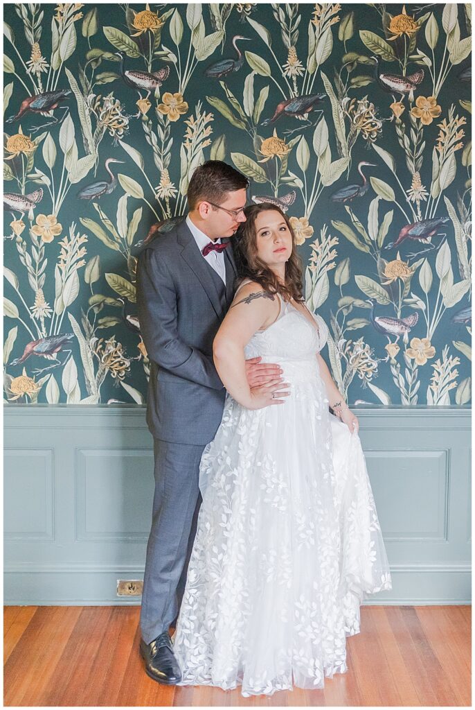 A bride and groom standing for a portrait in front of eclectic wall paper, the groom wearing a grey suit and the bride wearing a sleeveless, A-line gown with leafy lace pattern down the skirt.

Rust Manor House Wedding | Leesburg Wedding Photography | VA Wedding Venues